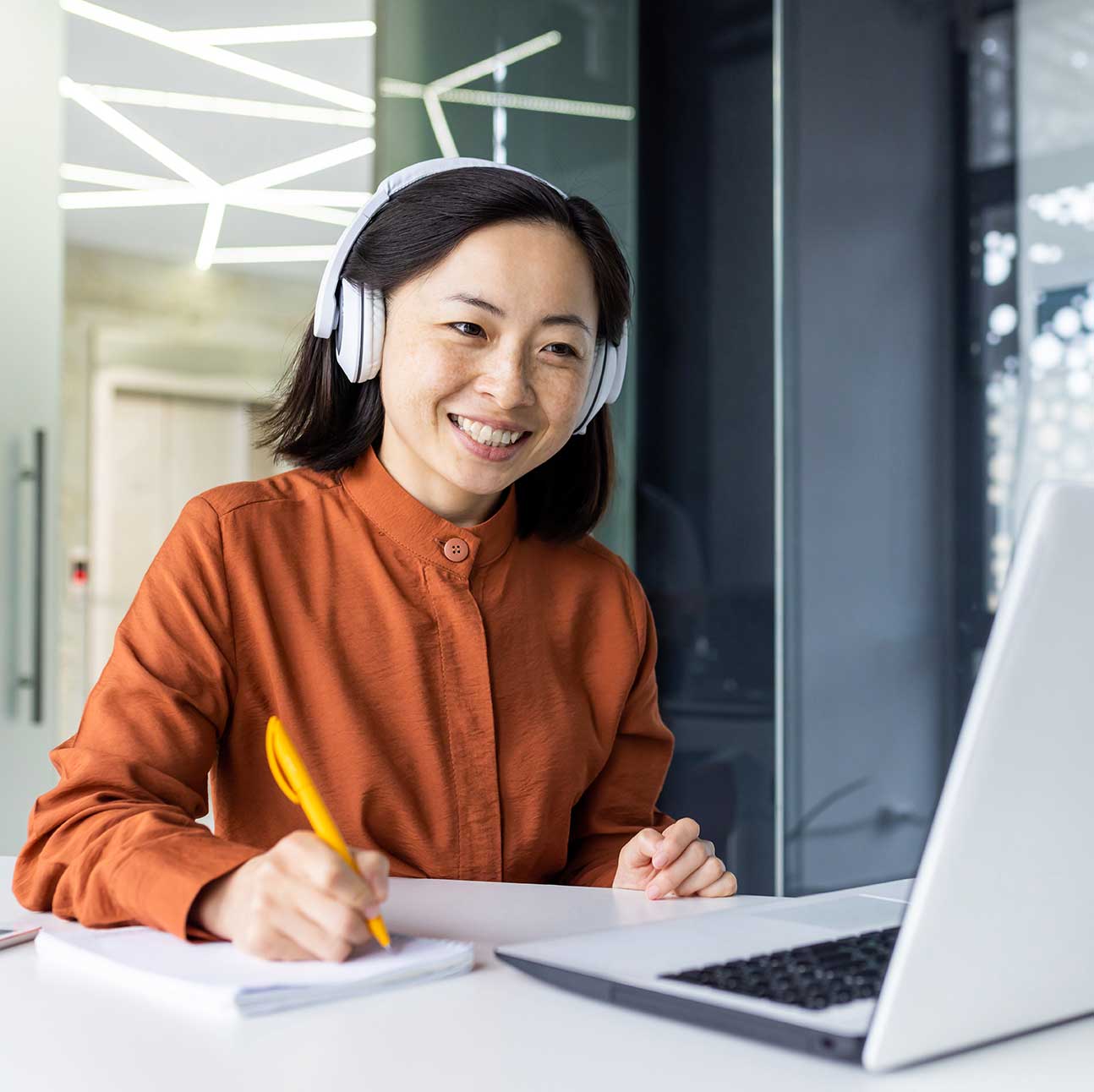 Business woman with headphones watching online training course at workplace, woman writing